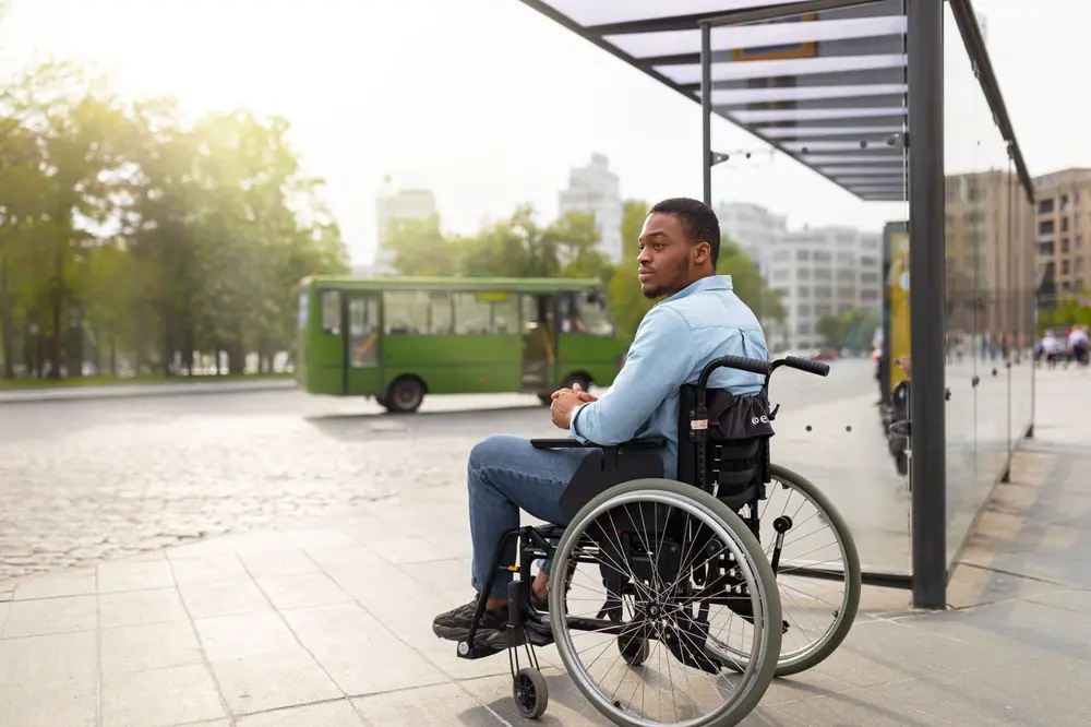 A man using a wheelchair waits at a bus stop as a bus pulls in.
