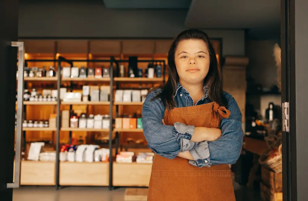 A young woman in a brown apron stands with her arms crossed and smiles in the doorway of a shop.